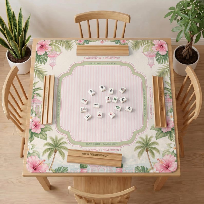 May include: A square mahjong mat featuring a cream-colored center with pink stripes, surrounded by a border of pink hibiscus flowers, green palm leaves, and pagoda illustrations. Four wooden tile racks are placed on the mat with mahjong tiles.