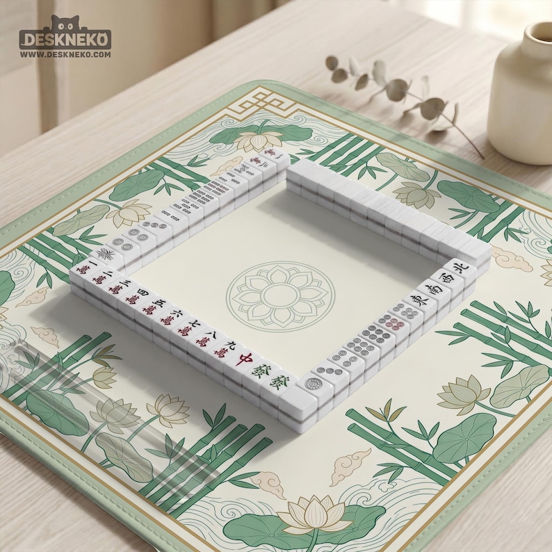 Puede incluir: Un juego de Mahjong completo dispuesto sobre un tapete decorativo. Las fichas blancas presentan caracteres rojos y negros. El tapete tiene un borde verde claro y un centro crema con dise&ntilde;os florales y de bamb&uacute;. El logotipo "DESKNEKO" es visible en la esquina superior izquierda.
