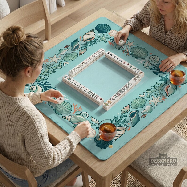 May include: A wooden square table with a light blue Mahjong mat decorated with seashells and coral. Two people are playing Mahjong, with tiles and drinks visible. The mat has the text "DESKNEKO" at the bottom.