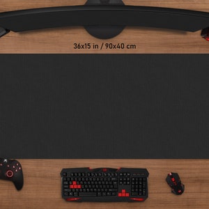 May include: A black rectangular gaming mouse pad, measuring 36 x 15 inches (90 x 40 cm), sits on a wooden surface. A black keyboard, mouse, controller, and headset with red accents are arranged around the pad. A curved monitor is above the pad.