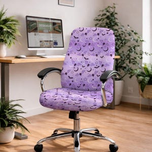 May include: A purple office chair with a Halloween-themed pattern of bats, moons, and stars. The chair has black armrests and a chrome base. The background includes a desk with a computer and plants.