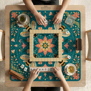May include: A square mahjong mat with a teal background featuring a central pink lotus flower, bees, and floral vines. The mat displays a square wall of ivory mahjong tiles, a tile rack with Chinese characters, two tea cups, and a small brown case.