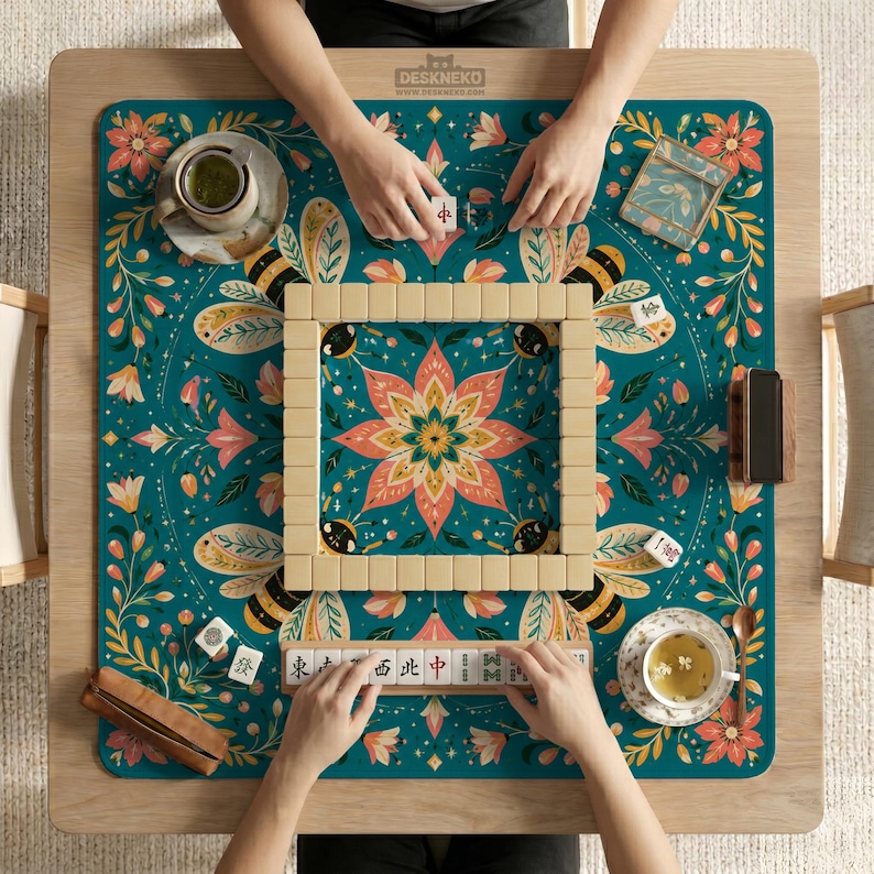 May include: A square mahjong mat with a teal background featuring a central pink lotus flower, bees, and floral vines. The mat displays a square wall of ivory mahjong tiles, a tile rack with Chinese characters, two tea cups, and a small brown case.