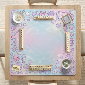 May include: A square mahjong game mat with a pastel ocean-themed design. The mat features illustrations of sea creatures, bows, and bubbles. Includes mahjong tiles, dice, and a small bowl with green balls. The text "DESKNEKO" is visible.