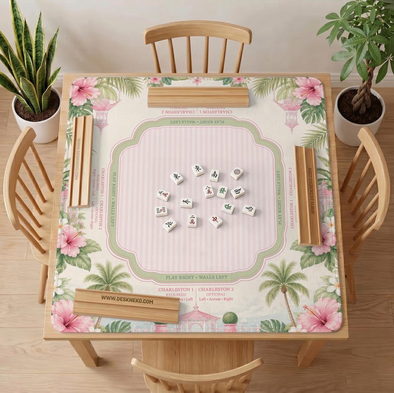 May include: A square mahjong mat featuring a light pink striped center, a green border, and a tropical floral pattern with palm trees and hibiscus flowers. Four wooden tile racks are placed on the edges. The mat includes text like DESKNEKO.COM.