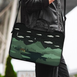 May include: A black laptop bag with a landscape design featuring a house, river, mountains, clouds, moon, and stars. The bag has a handle and a shoulder strap. The design is in shades of green and black.