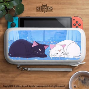 May include: A white and grey Nintendo Switch case with a cartoon illustration of two cats, one black and one white. The case is open, showing the Switch console. The text "DESKNEKO" is at the top.