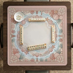May include: A square mahjong game board with a pink and white beach-themed design. The board features seashells, coral, and compass rose motifs. Wooden mahjong tiles and a small bowl with dice are on the board.