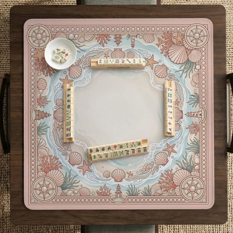 May include: A square mahjong game board with a pink and white beach-themed design. The board features seashells, coral, and compass rose motifs. Wooden mahjong tiles and a small bowl with dice are on the board.