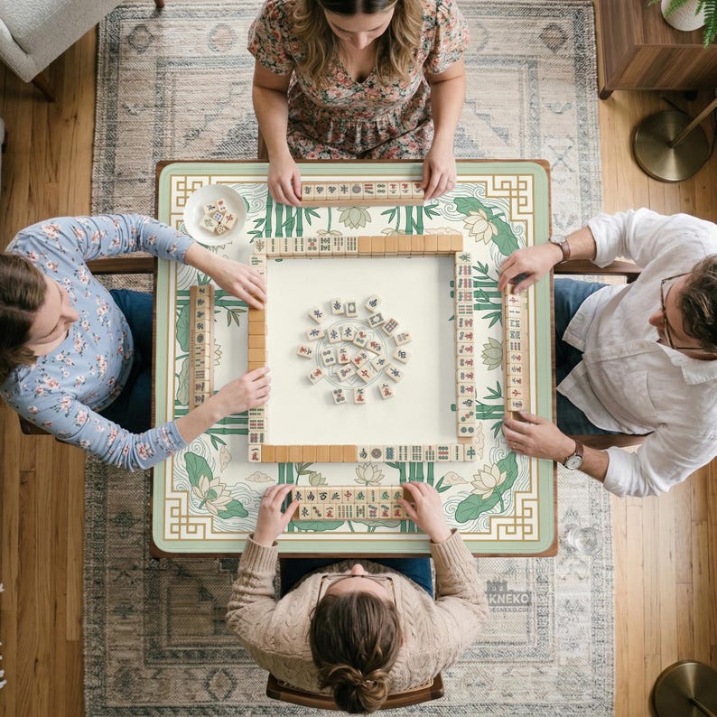 Puede incluir: Vista cenital de una partida de Mahjong en curso. La mesa tiene un dise&ntilde;o floral en verde claro y crema. Cuatro personas est&aacute;n sentadas alrededor de la mesa, cada una con una mano de fichas. Las fichas son beige con s&iacute;mbolos negros.