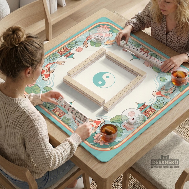 May include: A wooden mahjong table with a decorative mat featuring koi fish, pagodas, and a yin-yang symbol. The table is set for a game, with tiles arranged in a square and players holding racks of tiles. The mat has a teal border.