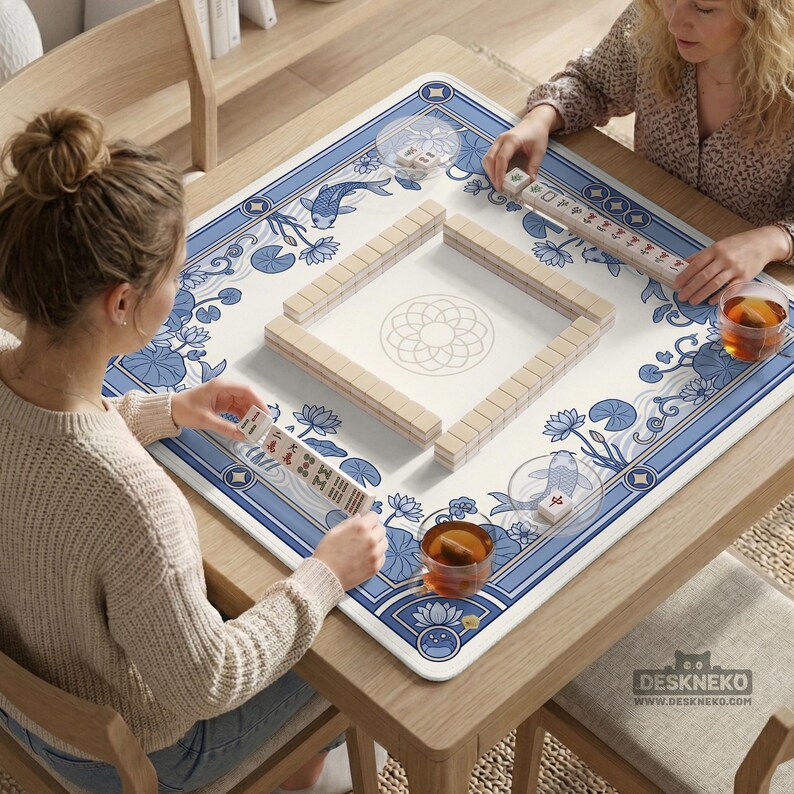 May include: A square Mahjong game table with a blue and white floral design. The table is set for a game, with tiles arranged in a square and players holding tiles. The table has a wooden frame and is set on a wooden table.