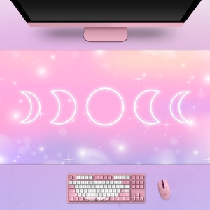 Puede incluir: Una alfombrilla de ratón rectangular grande con un fondo degradado en tonos pastel rosa, morado y azul. El diseño presenta un contorno blanco brillante de las fases lunares. También se ven un teclado, un ratón y unos auriculares rosas.