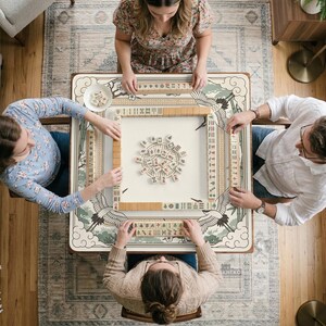 May include: Overhead view of four people playing Mahjong. The game is set on a square table with a cream-colored surface and a decorative border featuring cranes and floral designs. The players are seated around the table, each holding tiles.