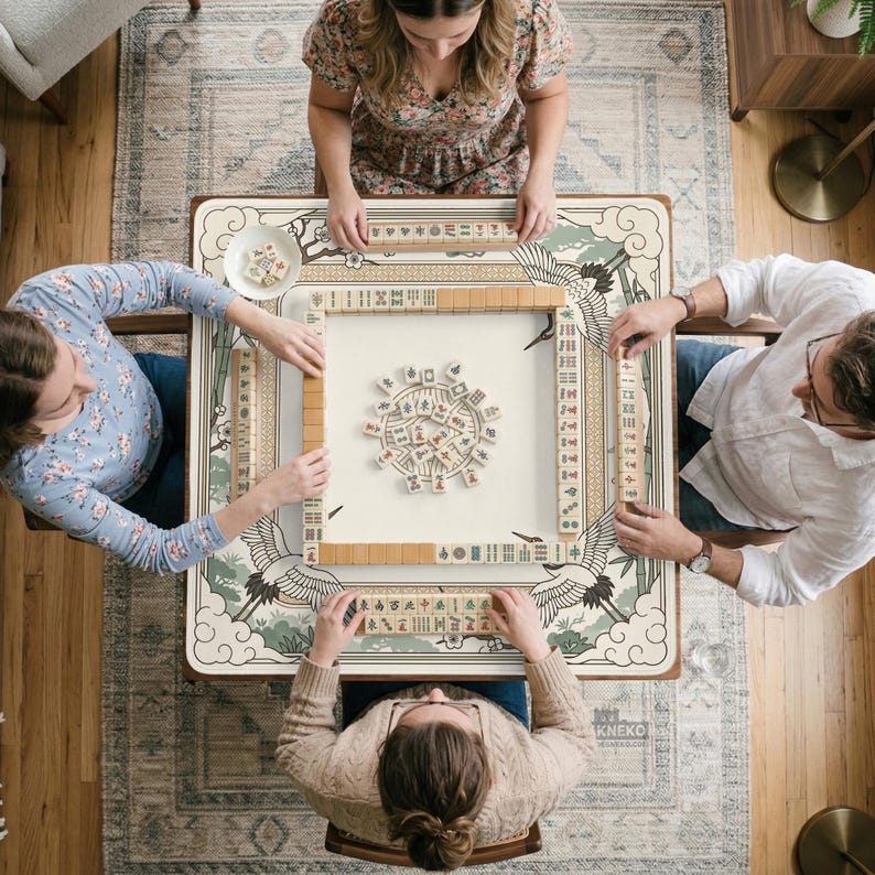 May include: Overhead view of four people playing Mahjong. The game is set on a square table with a cream-colored surface and a decorative border featuring cranes and floral designs. The players are seated around the table, each holding tiles.