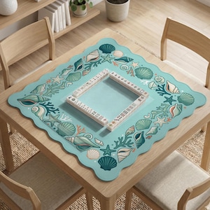 May include: A square, pale blue table mat with a scalloped edge, decorated with seashells, starfish, and coral. A wooden frame sits in the centre. The mat is on a wooden table.