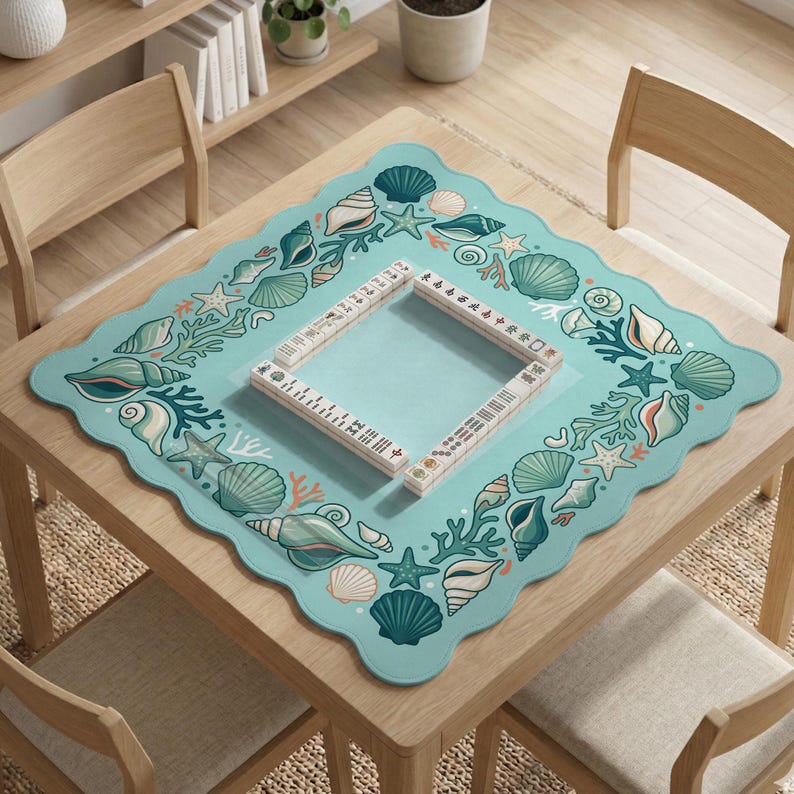 May include: A square, pale blue table mat with a scalloped edge, decorated with seashells, starfish, and coral. A wooden frame sits in the centre. The mat is on a wooden table.