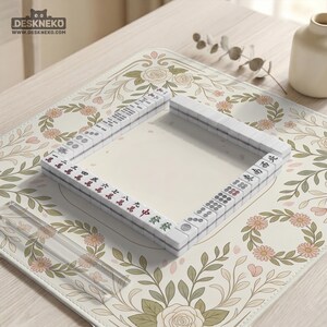 May include: A square mahjong tile set arranged in a hollow square formation on a cream-colored mat featuring a floral pattern of pink roses and green leaves. The white tiles display various black, red, and green Chinese characters and circular symbols.