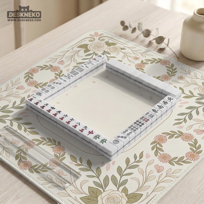 May include: A square mahjong tile set arranged in a hollow square formation on a cream-colored mat featuring a floral pattern of pink roses and green leaves. The white tiles display various black, red, and green Chinese characters and circular symbols.