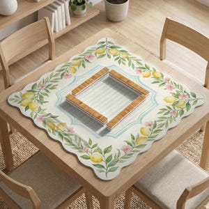 May include: A square, off-white tablecloth with a scalloped edge, adorned with watercolor lemons, pink flowers, and green leaves. A light blue border frames a striped center. Four wooden blocks are placed in the center of the tablecloth.