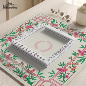 May include: A square mahjong mat featuring a cream-colored center with a pink circular brushstroke design. The border displays a pattern of pink cherry blossoms and green bamboo stalks. A square arrangement of white mahjong tiles sits in the center.