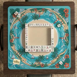 May include: A square mahjong game board with a tropical island design. The board has a teal border with illustrations of palm trees, sailboats, and flowers. White mahjong tiles are arranged in a square, ready for play.