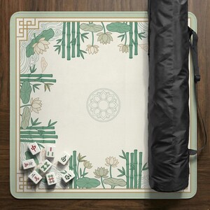 Puede incluir: Un tapete de juego de mahjong rectangular con una superficie color crema, adornado con bamb&uacute; verde, flores de loto y un dise&ntilde;o floral central. El tapete est&aacute; bordeado en verde claro e incluye una funda de transporte negra. Varias fichas de mahjong est&aacute;n esparcidas sobre el tapete.