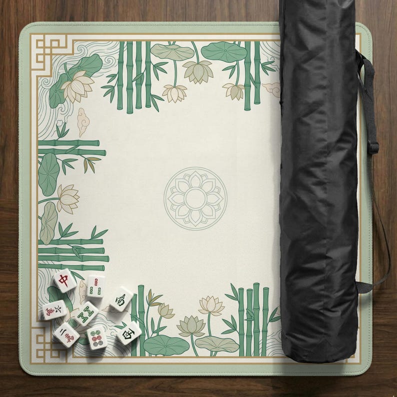 Puede incluir: Un tapete de juego de mahjong rectangular con una superficie color crema, adornado con bamb&uacute; verde, flores de loto y un dise&ntilde;o floral central. El tapete est&aacute; bordeado en verde claro e incluye una funda de transporte negra. Varias fichas de mahjong est&aacute;n esparcidas sobre el tapete.