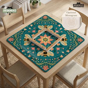 May include: A square desk mat featuring a teal background with a symmetrical pattern of bees, flowers, and foliage in shades of orange, yellow, and green. A wooden frame sits in the center, and a circular inset shows an optional monogram design.