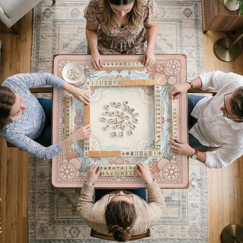 May include: A group of people playing Mahjong around a pink table with a sea-themed design. The table features coral, seashells, and a compass rose. Wooden tiles are arranged on the table, and players are seated around it.
