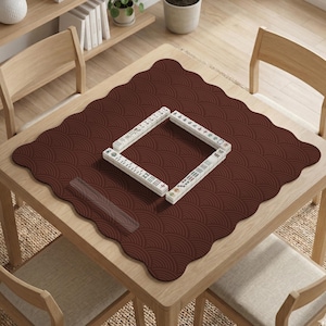 Puede incluir: Un tapete de mahjong marrón con borde festoneado y un patrón de abanico repetido. Las fichas de mahjong blancas están dispuestas en un cuadrado sobre el tapete. El tapete está sobre una mesa de madera clara.