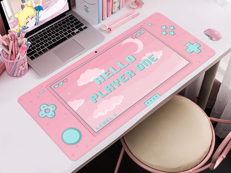 K&ouml;nnte beinhalten: Rosa Schreibtischunterlage mit einem Videospiel-Controller-Design. Die Matte zeigt den Text "HELLO PLAYER ONE" in pixeligen blauen Buchstaben sowie Wolken, Herzen und Sterne. Ein Laptop, Stifte und eine Maus sind ebenfalls zu sehen.