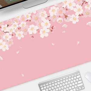 May include: A pink desk mat featuring a floral cherry blossom design. The blossoms are white and pink, with falling petals. A computer keyboard and mouse are visible at the bottom of the mat.