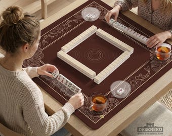 May include: A square wooden table with a brown Mahjong game mat featuring koi fish and decorative patterns. Beige Mahjong tiles are arranged in a square. Two people are playing the game, holding tiles. Clear glass cups with tea are on the table.