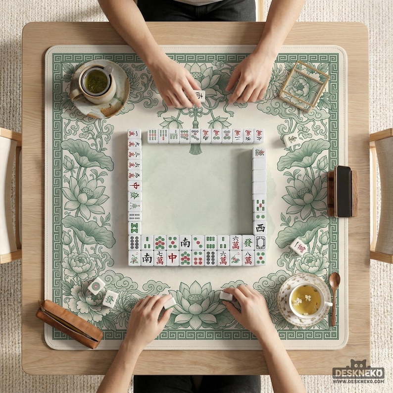 Puede incluir: Vista cenital de un juego de Mahjong en curso. El juego est&aacute; sobre una alfombra beige claro con un borde floral verde y dise&ntilde;os de flores de loto. Se ven las manos de los jugadores, colocando las fichas. Tazas de t&eacute; y accesorios tambi&eacute;n est&aacute;n sobre la mesa.