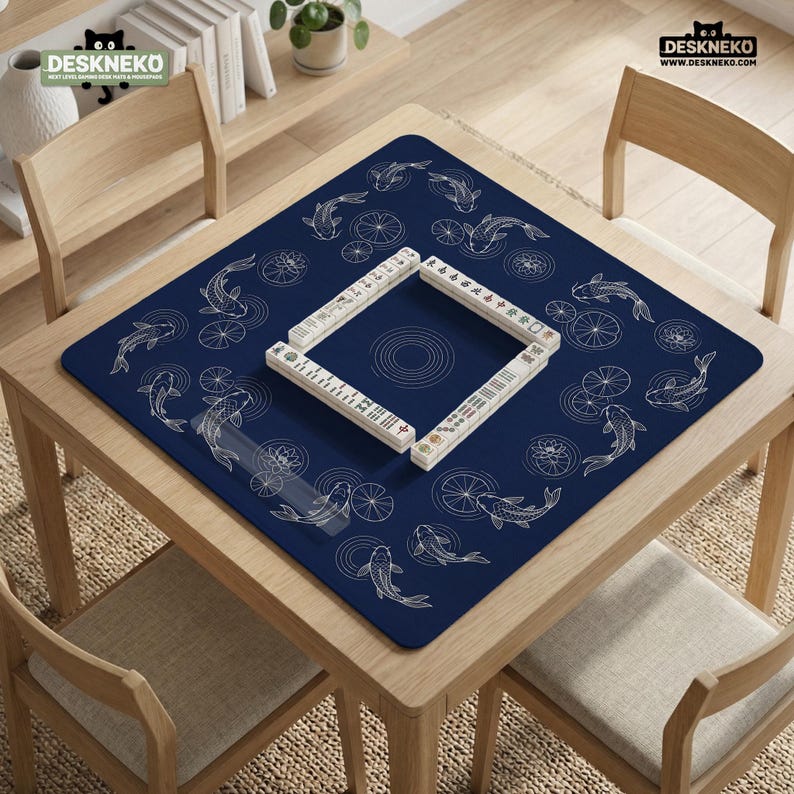 Puede incluir: Un tapete de mahjong azul marino con un dise&ntilde;o de peces koi y nen&uacute;fares. El tapete est&aacute; sobre una mesa de madera con cuatro sillas. Las fichas de mahjong est&aacute;n dispuestas en un cuadrado en el centro. La marca "DESKNEKO" es visible.