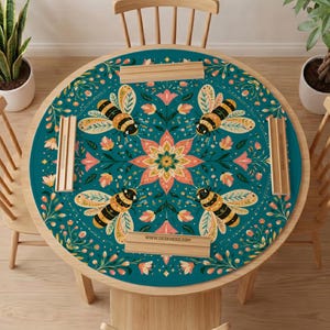 May include: A round teal table mat featuring a symmetrical pattern of four bees and a central floral motif in shades of pink, yellow, and green. The mat is accented with four light wood rectangular holders, each printed with the text WWW.DESKNEKO.COM.