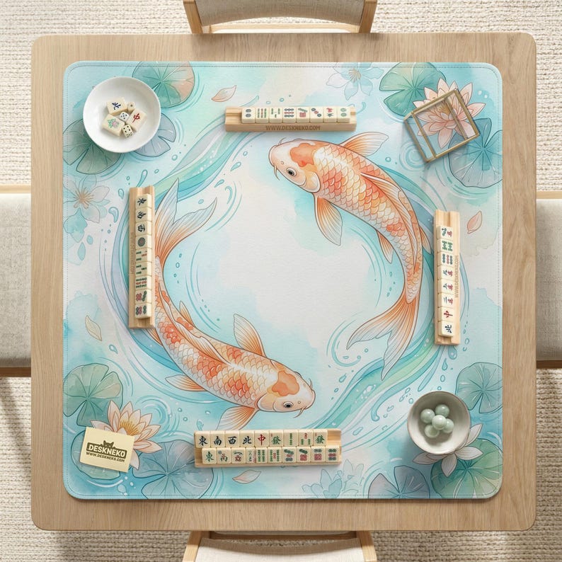 May include: A square mahjong mat featuring a light blue water background with two orange koi fish swimming in a circle, surrounded by lotus flowers and lily pads. Four wooden tile racks hold mahjong tiles, with extra tiles in a white bowl.