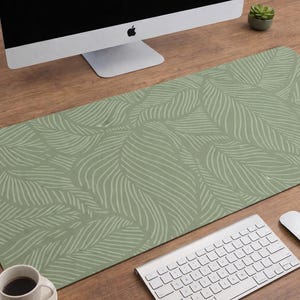 Op de afbeelding: Een rechthoekige bureaumat in saliegroen, met een herhalend patroon van lichtgroene gestileerde bladeren. De mat ligt op een houten bureau, met een computer, toetsenbord, muis en koffiekopje.