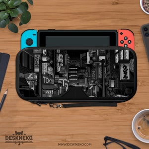 May include: A black and gray Nintendo Switch case featuring a pixelated Tokyo street scene. The case is on a wooden surface with a Switch console, a phone, and a cup of coffee. The case has a wrist strap and the text "TOKYO".