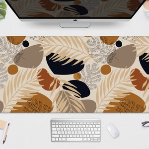May include: A rectangular desk mat featuring a beige background with a repeating pattern of abstract shapes and botanical elements in shades of brown, black, and tan. The mat is designed to protect a desk surface and provide a smooth surface for a mouse and keyboard.