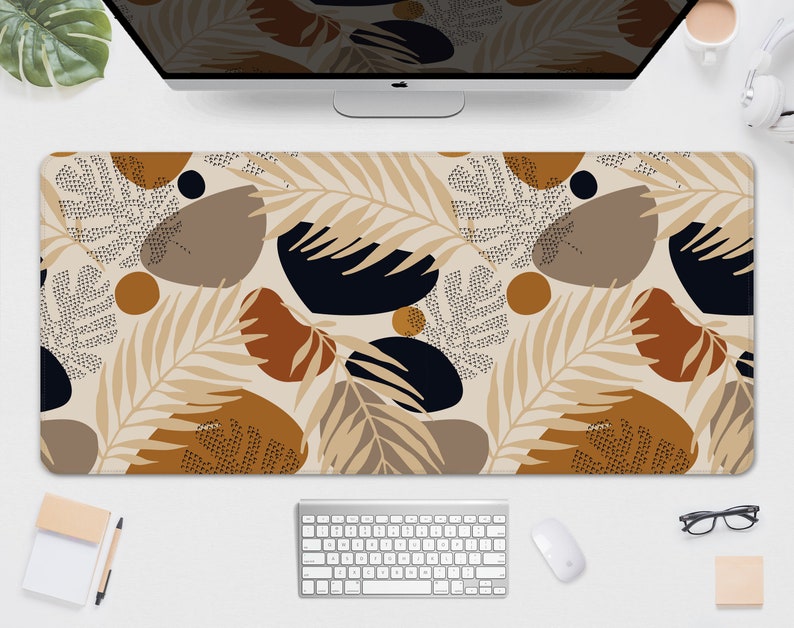May include: A rectangular desk mat featuring a beige background with a repeating pattern of abstract shapes and botanical elements in shades of brown, black, and tan. The mat is designed to protect a desk surface and provide a smooth surface for a mouse and keyboard.