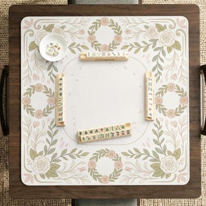 May include: A square mahjong mat featuring a white center and a border decorated with pink flowers, green leaves, and floral wreaths. The mat includes four wooden tile racks, a small white bowl with dice, and mahjong tiles arranged on the surface.