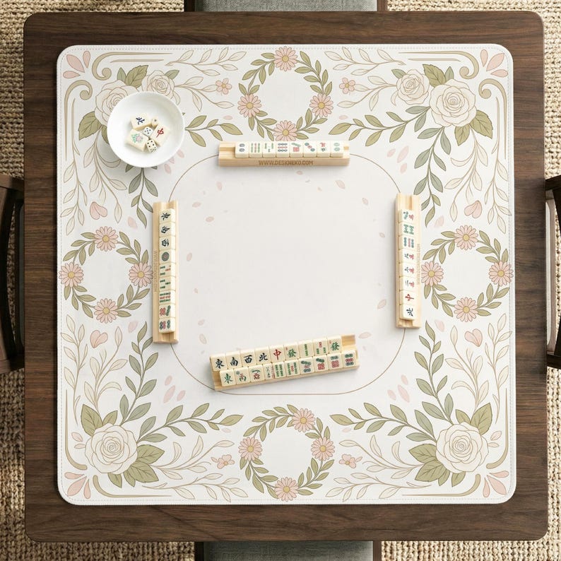 May include: A square mahjong mat featuring a white center and a border decorated with pink flowers, green leaves, and floral wreaths. The mat includes four wooden tile racks, a small white bowl with dice, and mahjong tiles arranged on the surface.