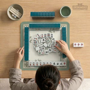 May include: A top-down view of a Mahjong game in progress. Teal tiles are arranged around a green mat with bamboo designs. White tiles with red and black symbols are scattered in the center. Accessories include a small bowl with dice and a cup of tea.