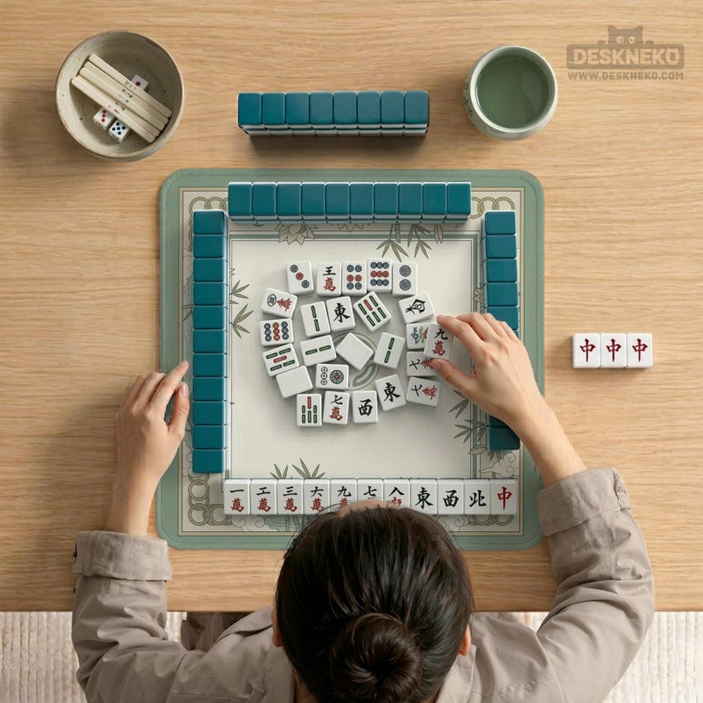 May include: A top-down view of a Mahjong game in progress. Teal tiles are arranged around a green mat with bamboo designs. White tiles with red and black symbols are scattered in the center. Accessories include a small bowl with dice and a cup of tea.