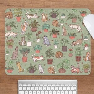 Puede incluir: Alfombrilla de rat&oacute;n rectangular con un fondo verde claro que presenta un patr&oacute;n de plantas en macetas y gatos juguetones en varias poses. Un rat&oacute;n blanco descansa en el lado derecho de la alfombrilla, con un teclado debajo.