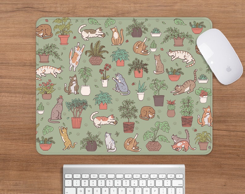 Puede incluir: Alfombrilla de rat&oacute;n rectangular con un fondo verde claro que presenta un patr&oacute;n de plantas en macetas y gatos juguetones en varias poses. Un rat&oacute;n blanco descansa en el lado derecho de la alfombrilla, con un teclado debajo.