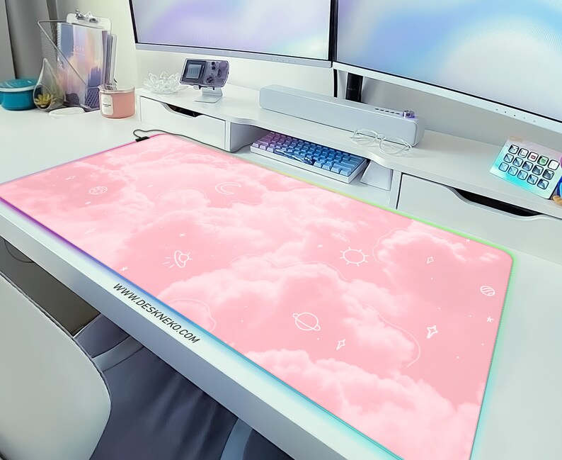 May include: A rectangular pink mouse pad with a cloud design and white celestial illustrations. The mouse pad has a colorful LED edge and the website address www.deskneko.com printed on the bottom left corner.