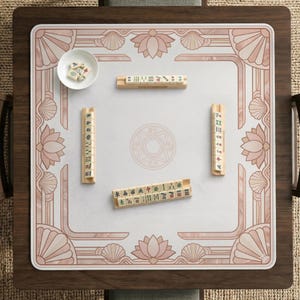 May include: A square mahjong table with a white playing surface and a decorative pink border featuring lotus flowers and seashells. The table includes mahjong tiles and a small bowl with dice.